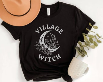 Puede incluir: Camiseta negra con un gráfico blanco de una luna creciente con cristales y flores. El texto "Village Witch" está impreso debajo de la luna.