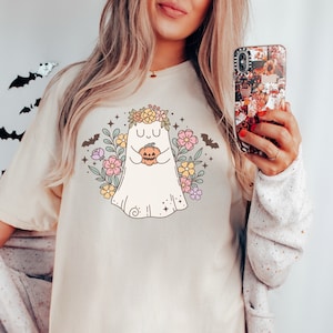 May include: A white t-shirt with a cute cartoon ghost wearing a flower crown and holding a pumpkin. The ghost is surrounded by flowers and bats.