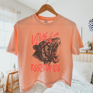 May include: A peach-colored cropped t-shirt with a graphic of a roaring tiger and the text "Vive La Resistance" in red.