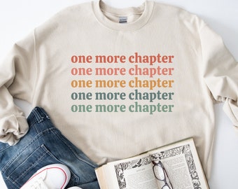 May include: A beige sweatshirt with the text "one more chapter" repeated five times in different colors, including red, orange, yellow, and green.