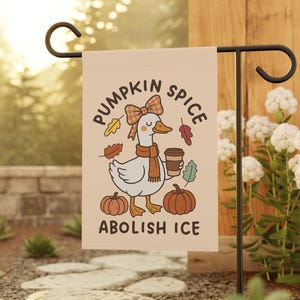May include: A beige garden flag featuring a cartoon goose with a bow, scarf, and coffee cup, surrounded by pumpkins and leaves. Text reads "PUMPKIN SPICE" and "ABOLISH ICE". The flag is displayed on a black metal stand in a garden setting.