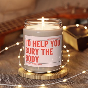 May include: A brown soy candle with a white label that reads "I'd help you bury the body" and "Everything After Company" in smaller text. The candle is sitting on a wooden coaster.