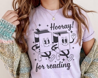 May include: A light purple t-shirt with a graphic of two cartoon books holding hands and the text "Hooray! for reading".