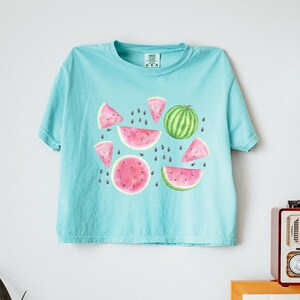 May include: A light blue t-shirt featuring a watercolor design of watermelon slices and a whole watermelon. The design includes pink watermelon slices with black seeds and a green watermelon with dark green stripes. The shirt is hanging on a white wall.