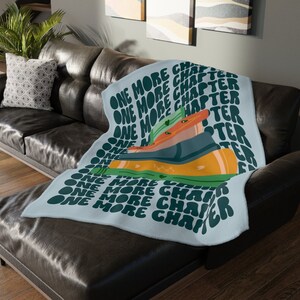 May include: A soft blue throw blanket with the text "One More Chapter" repeated in a green and white pattern. The blanket features a stack of books with a green, orange, and blue design.