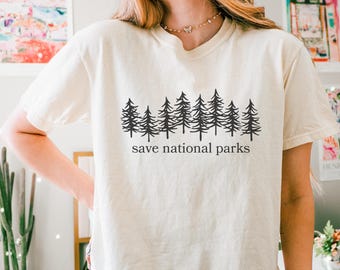 May include: Cream-colored t-shirt with a black graphic of pine trees and the text "save national parks." The shirt has short sleeves and a crew neck. The model is wearing dark brown leggings.