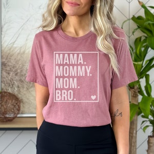 May include: A mauve t-shirt with a white box and the text "MAMA. MOMMY. MOM. BRO." inside the box. A small white heart is in the bottom right corner of the box.