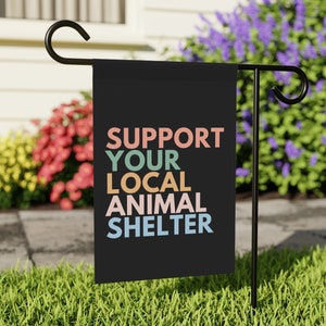 May include: A black garden flag with the text "SUPPORT YOUR LOCAL ANIMAL SHELTER" in colorful letters. The flag is displayed on a black metal garden flag stand.