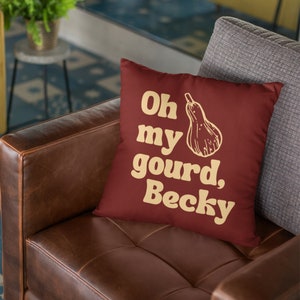 May include: A square throw cushion in a deep maroon colour featuring a light yellow graphic of a gourd and the text "Oh my gourd, Becky" printed in a bold, rounded font on the front surface of the cushion fabric.