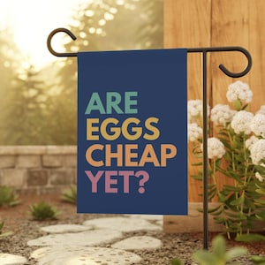 May include: A navy blue garden flag with the text "ARE EGGS CHEAP YET?" in colorful letters. The flag is hanging on a black metal stand, set in a garden with stone pavers and greenery.