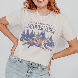 Può includere: T-shirt color crema con la scritta "MAY OUR DAUGHTERS BE UNGOVERNABLE" sopra una grafica di una persona che cavalca una tigre attraverso una foresta. La grafica è nei toni dell'arancione, blu e marrone.