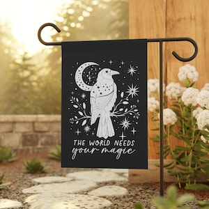 May include: Black garden flag with a white silhouette of a crow perched on a branch with a crescent moon and stars. The text "The world needs your magic" is printed below the crow.
