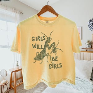 May include: Yellow t-shirt with the text "GIRLS WILL BE GIRLS" and a green illustration of two praying mantises. The shirt is hanging on a wooden hanger.
