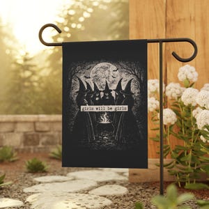 May include: A black garden flag featuring a white graphic illustration of five figures in pointed hats standing in a circle around a cauldron under a full moon. The text "girls will be girls" is printed in a white rectangular box in the center.