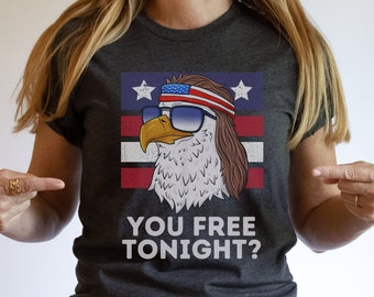 Puede incluir: Una camiseta gris oscuro con un gráfico de un águila con gafas de sol y un pañuelo rojo, blanco y azul. El águila está frente a un fondo a rayas rojo, blanco y azul. El texto "You free tonight?" está impreso debajo del águila.