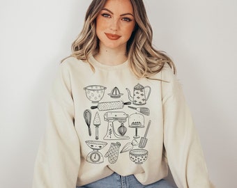 May include: Cream-colored sweatshirt featuring a black line drawing of various kitchen tools. The design includes a colander, rolling pin, whisk, mixer, and other baking essentials. The overall theme is centered around baking and cooking.