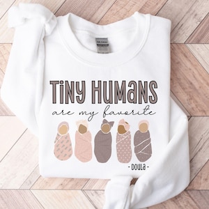 May include: A white long-sleeve sweatshirt featuring the text "Tiny Humans are my favourite" in dark brown and black lettering. Below the text are five illustrations of swaddled infants in various shades of pink and brown, with "- DOULA -" below.
