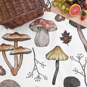 Może przedstawiać: Biały koc piknikowy z fantazyjnym wzorem grzyb&oacute;w. Koc przedstawia r&oacute;żne ilustracje grzyb&oacute;w w odcieniach brązu, beżu i czerwieni. Widoczny jest r&oacute;wnież wiklinowy kosz piknikowy i drewniana deska do krojenia z owocami i winogronami.