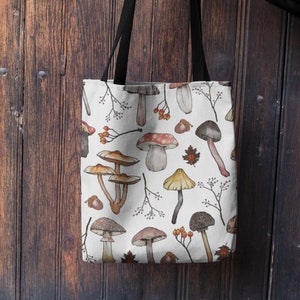 May include: A white canvas tote bag with black straps features a repeating pattern of watercolor mushrooms, leaves, and berries.