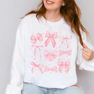 May include: White sweatshirt with a pink watercolor design of various bows and hearts.