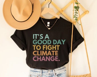 May include: Black t-shirt with the text "It's a good day to fight climate change." in rainbow colors. The shirt is hanging on a wooden hanger with a brown hat and a straw bag.