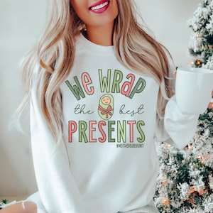 May include: White sweatshirt with green and pink text that reads "We wrap the best presents #motherbabyunit". The text is surrounded by a green and pink graphic of a baby wrapped in a blanket with a red bow.