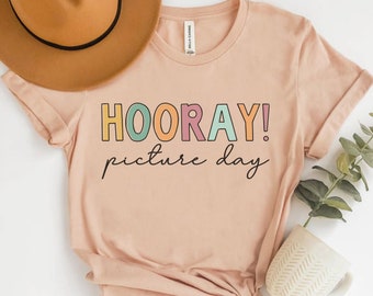 May include: A light pink t-shirt with the text "HOORAY! picture day" in colorful letters. The letters are outlined in black and have a slight shadow effect.