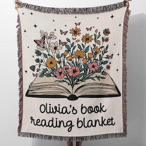May include: A woven throw blanket with a cream background and a colorful floral design. The flowers are growing out of an open book with the text "Olivia's book reading blanket".