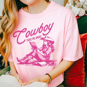 Peut inclure: Un t-shirt rose clair avec le mot "Cowboy" en &eacute;criture cursive et l'expression "Take Me Away" imprim&eacute;e en dessous. Le t-shirt pr&eacute;sente un graphisme d'une femme portant un chapeau de cowboy lisant un livre. Le t-shirt est en mati&egrave;re douce.