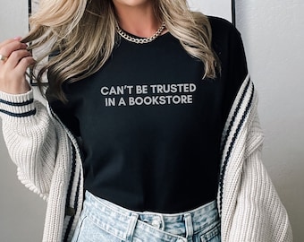 Puede incluir: Una camiseta negra con el texto "CAN'T BE TRUSTED IN A BOOKSTORE" impreso en la parte delantera. La camiseta la lleva puesta una persona con el pelo largo y rubio y un collar de cadena dorada.