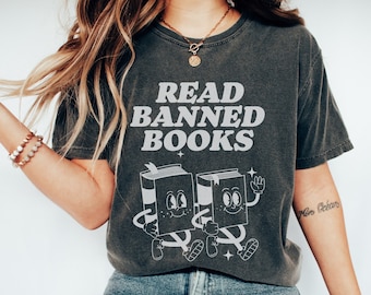 May include: A dark gray t-shirt with the text "READ BANNED BOOKS" in white. The shirt features a cartoon illustration of two books with legs and faces, walking together.
