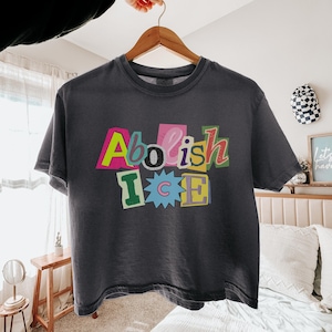 May include: Dark gray t-shirt with the words "Abolish ICE" in colorful, cut-out style letters. The shirt is hanging on a wooden hanger, with a hand holding the hanger. The shirt is a casual, unisex style.