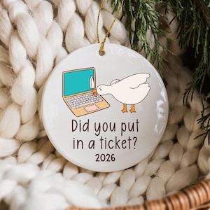 May include: A round white ceramic ornament featuring a graphic of a white goose holding a knife while looking at a laptop. The text reads Did you put in a ticket? and 2026. A thin gold string is attached to the top for hanging purposes.