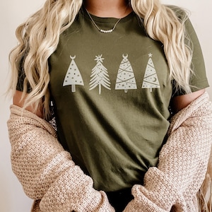 May include: A green t-shirt with a white graphic of five stylized Christmas trees. The trees have different designs, including stars, moons, and crescent moons. The person wearing the shirt is also wearing a beige cardigan.