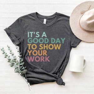 May include: A dark gray t-shirt with the text "It's a good day to show your work" in rainbow colors. The text is stacked vertically with each word in a different color.