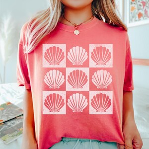 May include: A pink t-shirt with a white graphic of nine seashells arranged in a grid pattern.