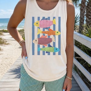 May include: Cream-colored tank top featuring a colorful fish design. The graphic includes four fish with various patterns and colors, set against blue vertical stripes. The model is also wearing green and white checkered shorts.