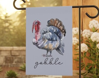 May include: A light gray garden flag featuring a watercolor illustration of a turkey. The turkey has a red wattle and a brown and blue feathered body. The word "gobble" is written in a script font below the turkey.