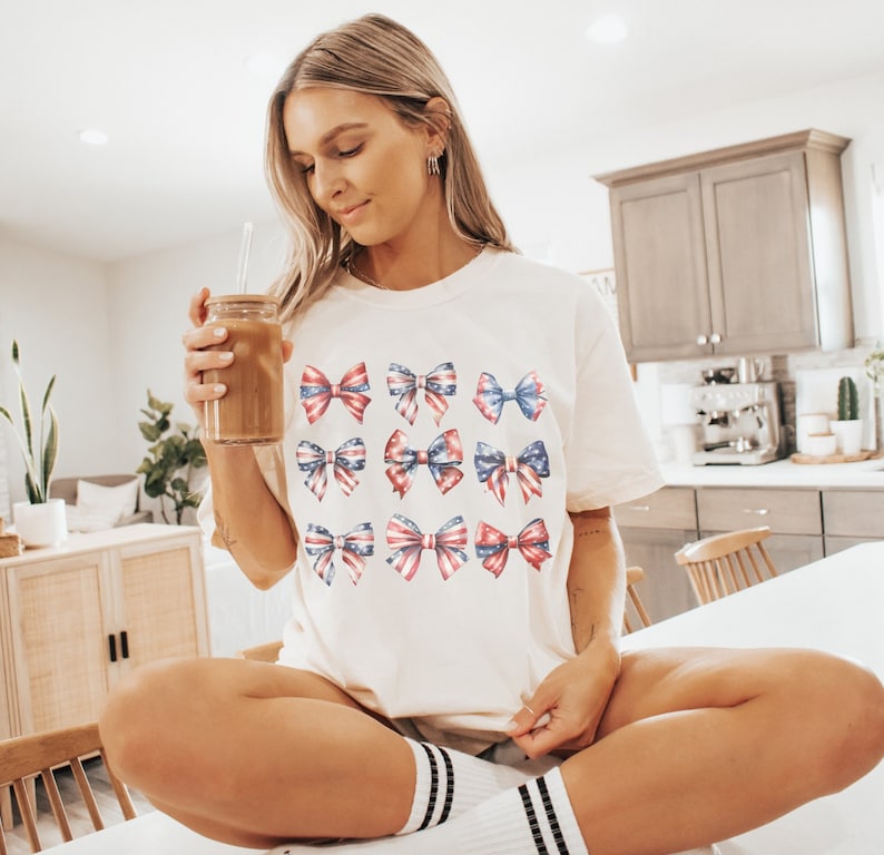 May include: A white t-shirt with a pattern of red, white, and blue bows. The bows are arranged in a grid pattern and are reminiscent of the American flag. The shirt is being worn by a person holding a glass of iced coffee.