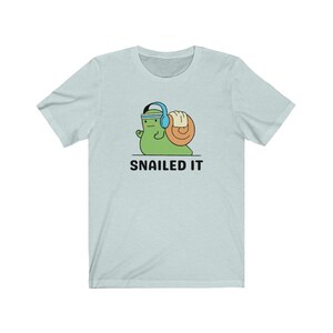 K&ouml;nnte beinhalten: Ein hellblaues T-Shirt mit einer Cartoon-Schnecke, die Kopfh&ouml;rer tr&auml;gt und ein Gewicht hebt. Der Text "SNAILED IT" ist unter der Schnecke aufgedruckt.