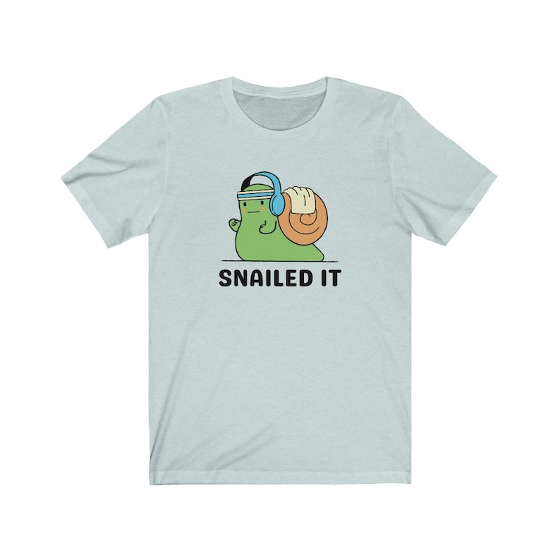 K&ouml;nnte beinhalten: Ein hellblaues T-Shirt mit einer Cartoon-Schnecke, die Kopfh&ouml;rer tr&auml;gt und ein Gewicht hebt. Der Text "SNAILED IT" ist unter der Schnecke aufgedruckt.