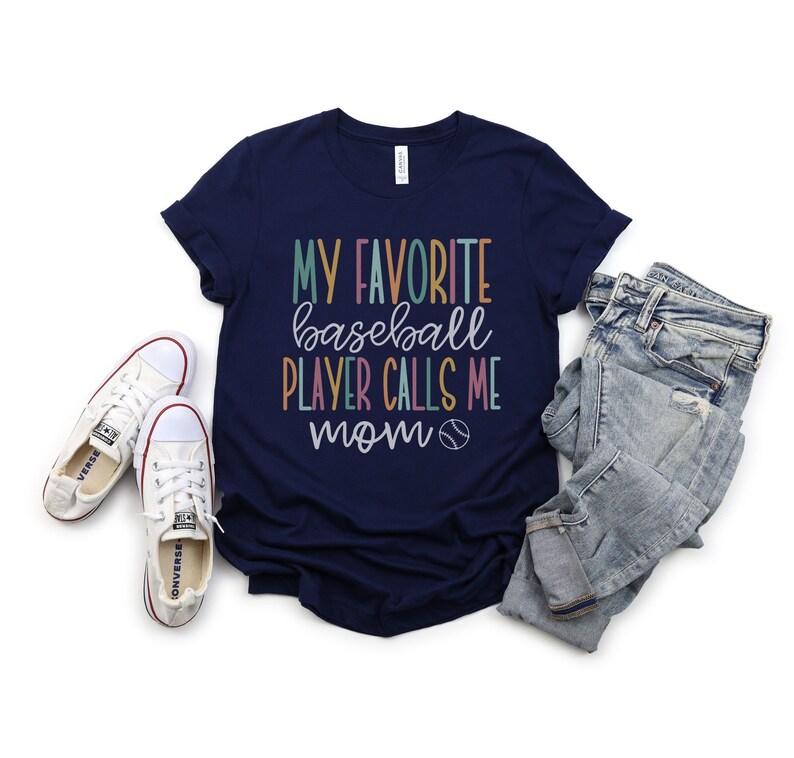 Op de afbeelding: Marineblauw T-shirt met de tekst "MY FAVORITE baseball PLAYER CALLS ME mom" in kleurrijke letters. Het shirt wordt gecombineerd met een lichtblauwe jeans en witte canvas sneakers.