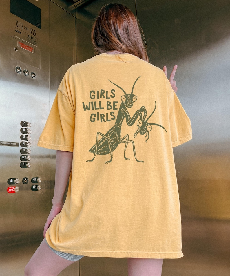 May include: Mustard yellow t-shirt with a green graphic of two praying mantises and the text "GIRLS WILL BE GIRLS". The shirt has short sleeves and a relaxed fit, suitable for casual wear.