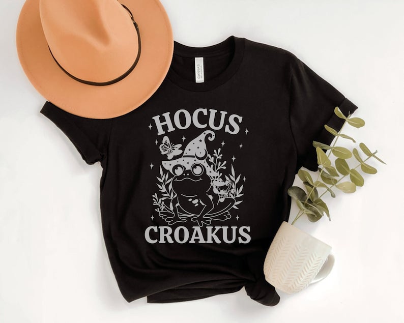 May include: Black t-shirt with a white graphic of a frog wearing a witch hat and the text "Hocus Croakus".