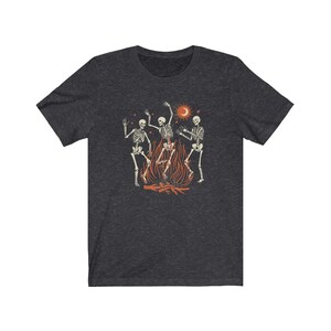 May include: A dark heather grey t-shirt with a graphic of three skeletons dancing around a campfire. The skeletons are white and the campfire is orange. There is a crescent moon and a sun in the background.