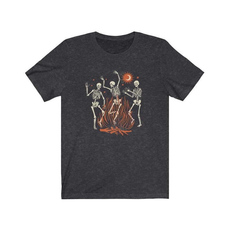 May include: A dark heather grey t-shirt with a graphic of three skeletons dancing around a campfire. The skeletons are white and the campfire is orange. There is a crescent moon and a sun in the background.
