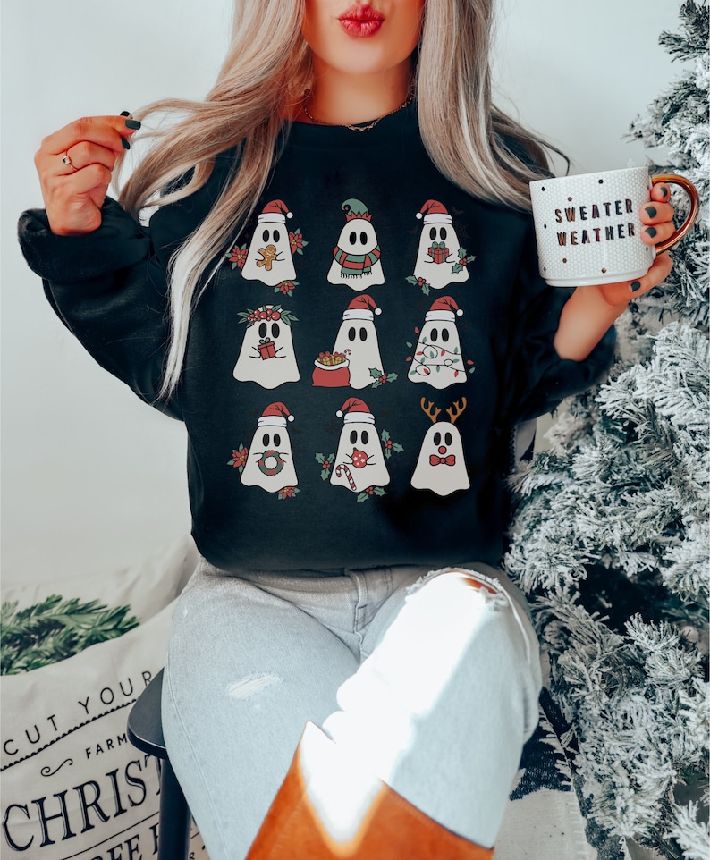 Op de afbeelding: Zwart sweatshirt met een kerstgeestontwerp. De geesten dragen kerstmanmutsen, elfenmutsen, rendiergewei en andere feestelijke accessoires. De tekst "Sweater Weather" staat op een witte mok.