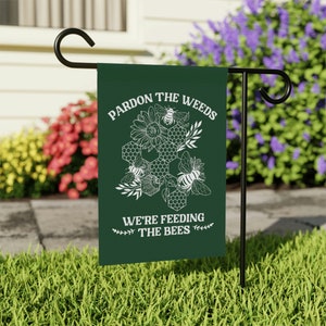 May include: A green garden flag with white text that reads "Pardon the weeds, we're feeding the bees". The flag features a white illustration of a honeycomb with bees and sunflowers.