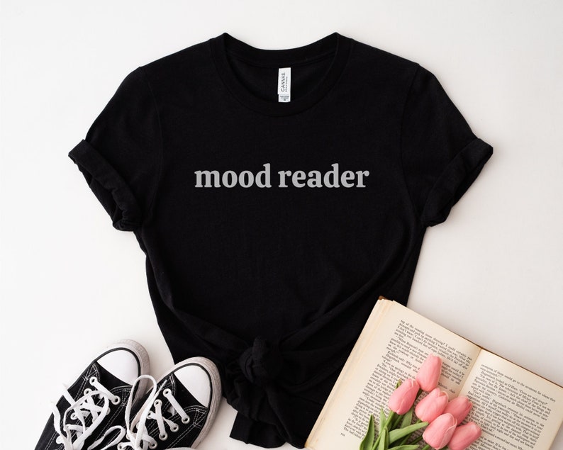 May include: Black t-shirt with the text "mood reader" in white lettering. The shirt is laying on a white surface with a book and flowers.