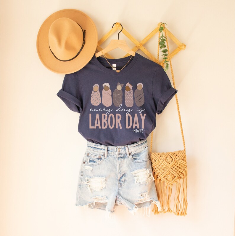 Op de afbeelding: Een marineblauw t-shirt met een afbeelding van vijf baby's in doeken en de tekst "every day is LABOR DAY - MIDWIFE - "